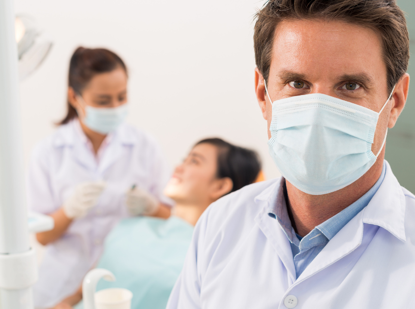 Dentist wearing a mask standing in front of camera while another dentist is in the back with a patient.
